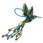 A handmade beaded hummingbird ornament, featuring vibrant blue, green, and gold seed beads with dangling bead strands and faceted bronze beads, set against a plain white background.