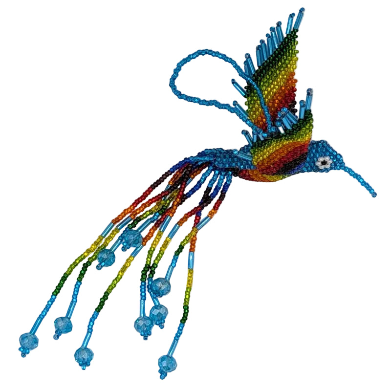 A colorful beaded hummingbird ornament with vibrant rainbow-colored wings and tail feathers, light blue body, and a small white-and-black eye, photographed on a plain white background.