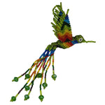 A handmade beaded hummingbird ornament, featuring a vibrant rainbow body and wings with dangling rainbow beaded tails with green crystals, photographed on a plain white background.